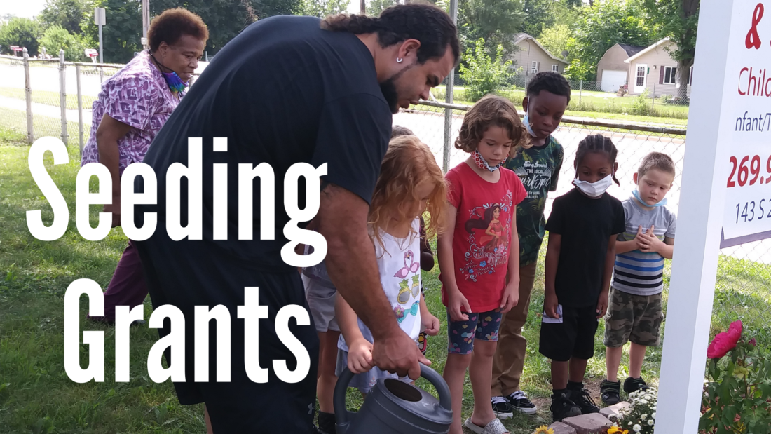 Image shows white text that reads Seeding Grants over a photo of a parent helping a group of small children water plants.